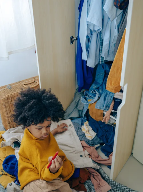 Child in yellow jumper with messy wardrobe, holding red toy; cluttered bedroom scene.
